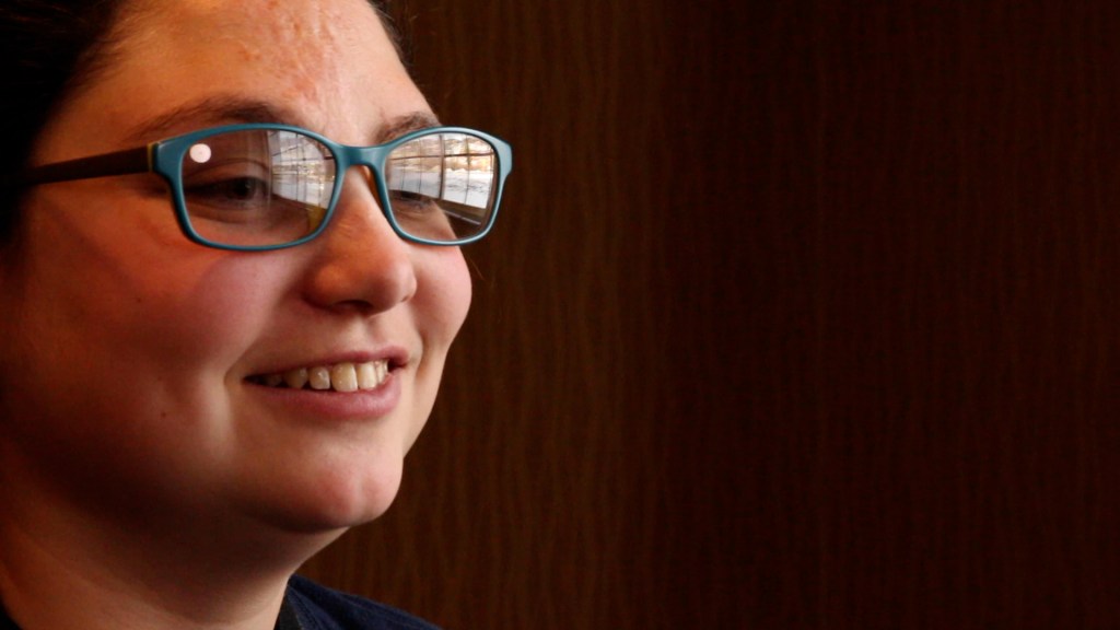 A woman wearing light blue glasses smiles into the camera. This is a still from her film interview.