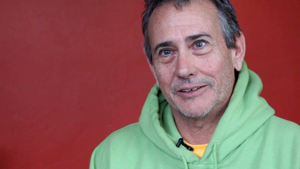 A white man with short brown hair with white strips looks intently into the camera. This is a still from his film interview.