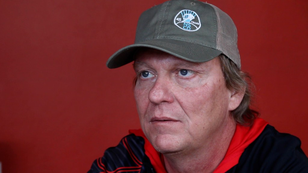 A man wearing a green hat looks into the camera. This is a still from his film interview.